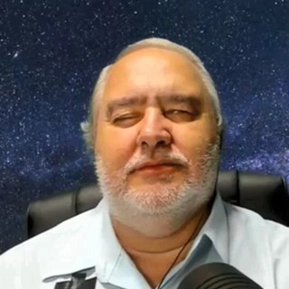 Francisco Lima is a white man, he has silver hair and he wears a white shirt. He is sits on a leather chair and he speaks at a microphone. Behind him, a green chromakey shows the space and the stars.